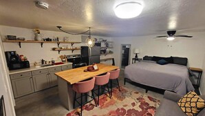 Ein kompaktes Hotelzimmer mit Küchenzeile, einem Bett, einer kleinen Sitzecke und Deckenventilator.