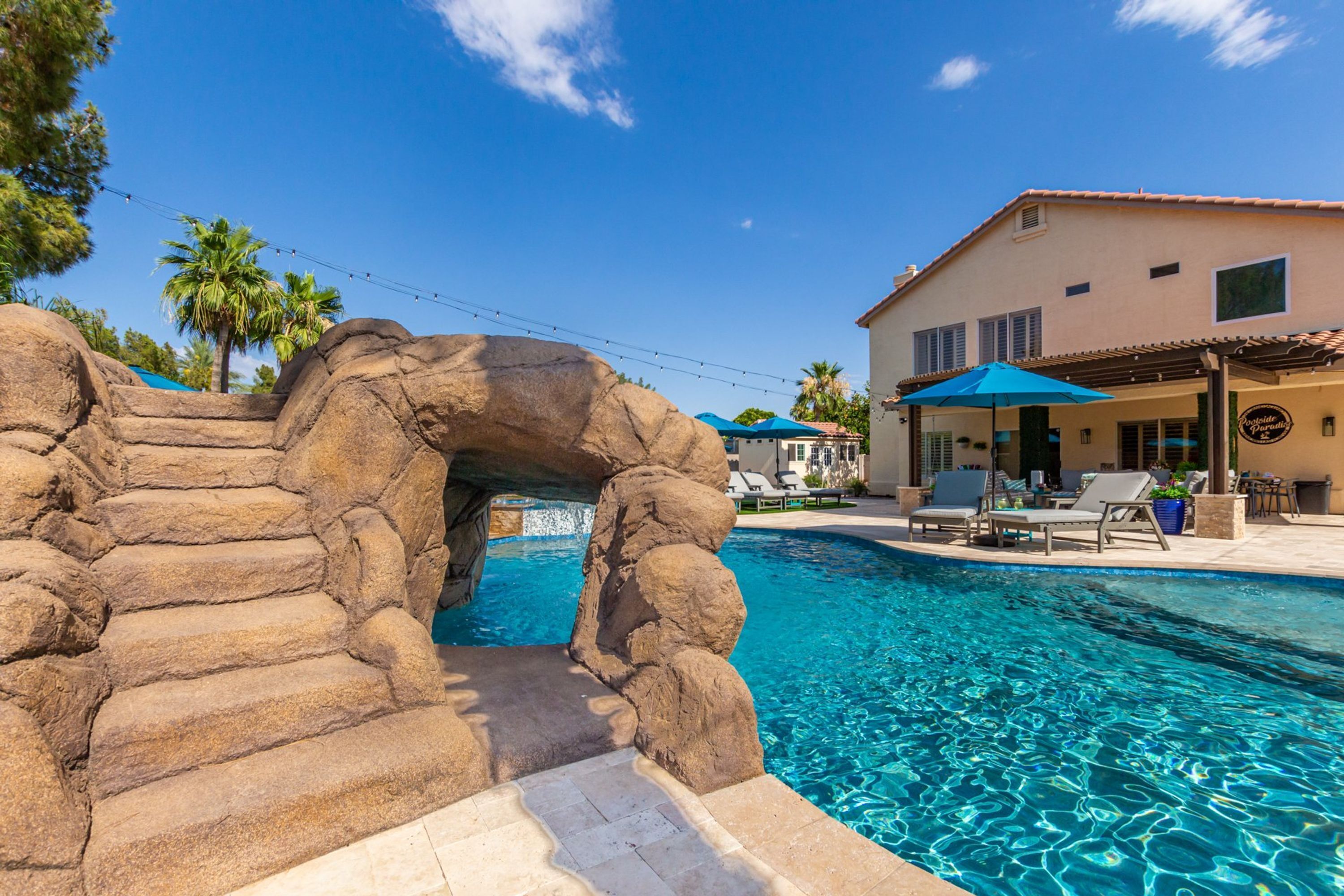 A pool area with a rock arch entrance, lounge chairs, and a building with a blue awning.