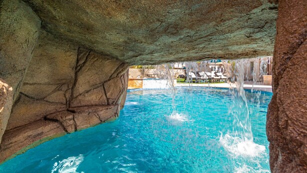 Ein Pool mit klarem, blauem Wasser, einem Wasserfall und Blick auf einen Resortbereich mit Gebäuden und einem Poolbereich.