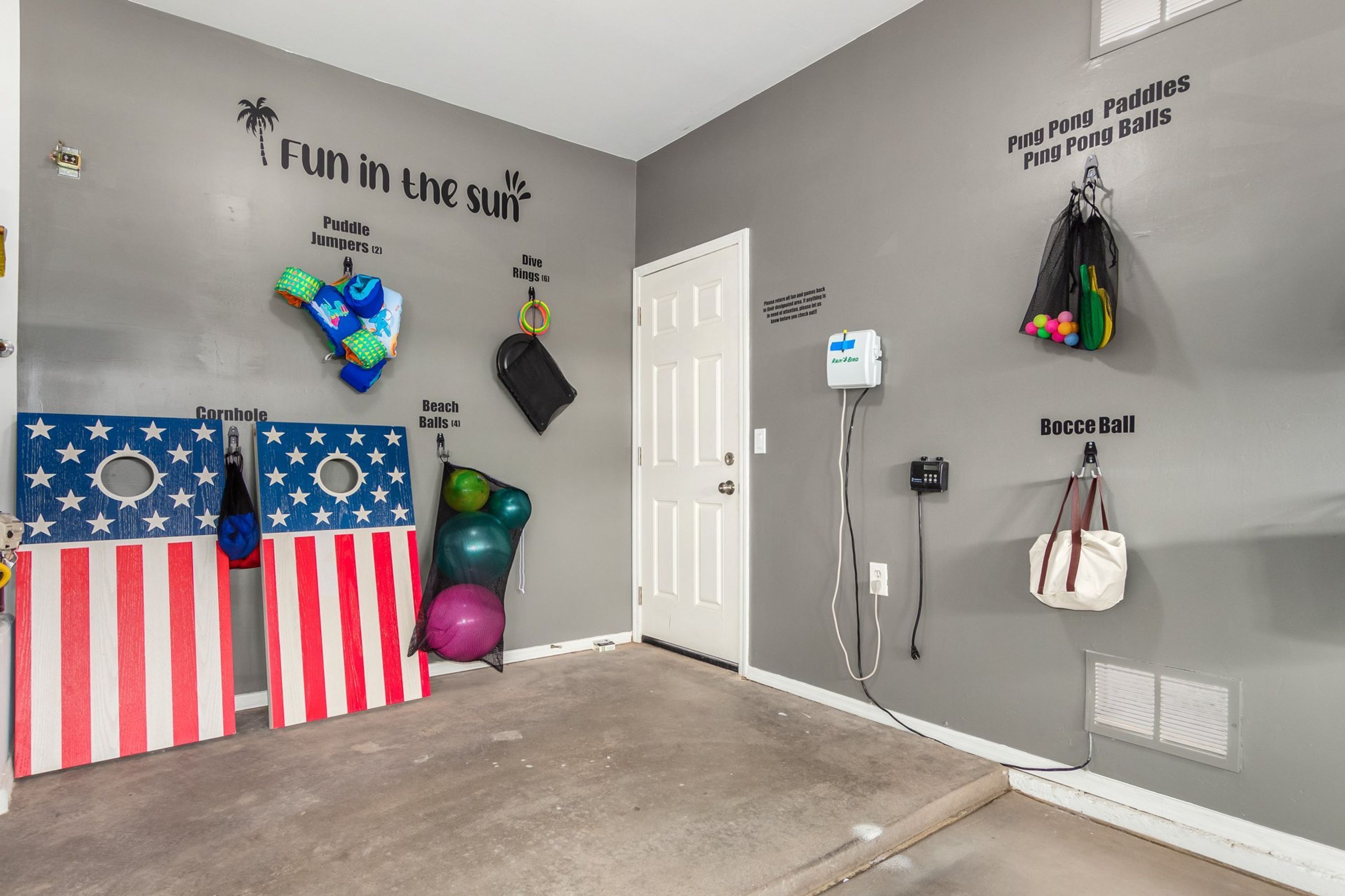 Quarto com parede cinzenta exibindo diversos equipamentos esportivos como raquetes, bolas e elásticos. Há uma porta branca, uma pequena janela e uma tomada na parede.