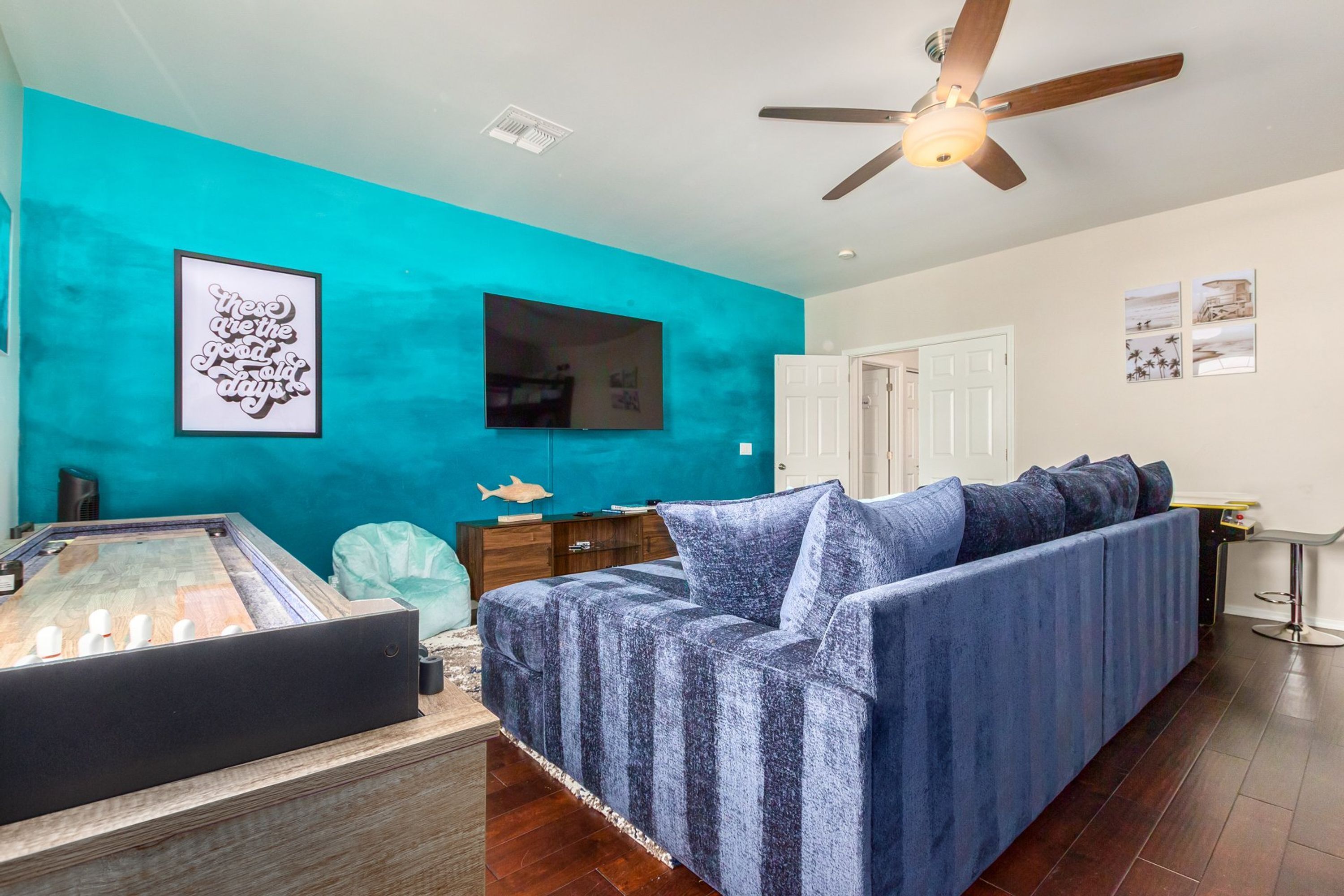 A room with a blue sofa, a foosball table, a ceiling fan, and a TV mounted on the wall.