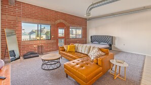 Ein modernes Schlafzimmer mit Backsteinwand, großem Fenster, einer Couch, einem runden Couchtisch und einem Bett mit grauem Kopfteil.
