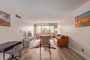 A modern living room with a wooden floor, a desk, a rocking chair, a sofa, and a large window offering a view of the outdoors.