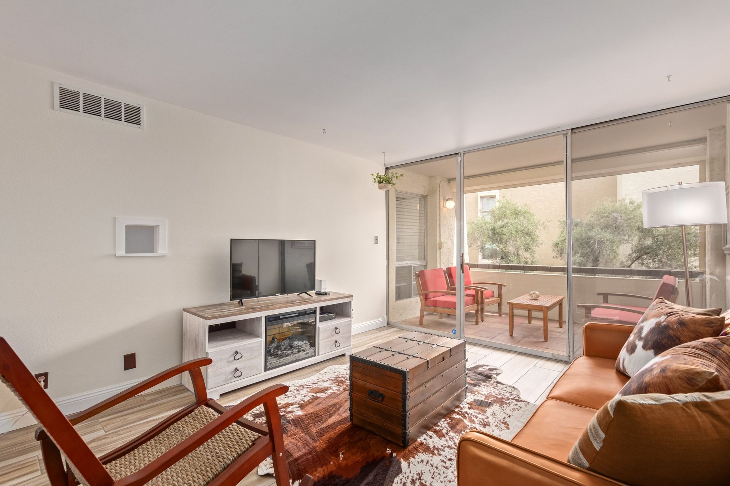 A modern living room with a sofa, a wooden coffee table, a TV on a white cabinet, and a sliding glass door leading to an outdoor patio with red chairs.