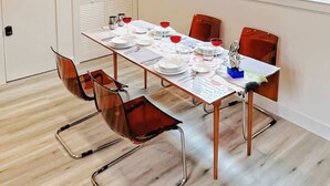 Un coin repas avec une longue table dressée pour quatre personnes, entourée de quatre chaises, une fenêtre avec des stores et une porte dotée d’une poignée.