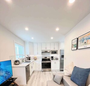 Una cocina moderna con armarios blancos, electrodomésticos de acero inoxidable y un horno empotrado. Hay una zona de comedor con un sofá de color claro y un televisor sobre un mueble.