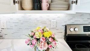 Une cuisine avec un meuble blanc contenant des verres transparents, des tasses noires et roses, et des assiettes empilées. Un panier en osier rempli de fleurs est posé sur le comptoir.