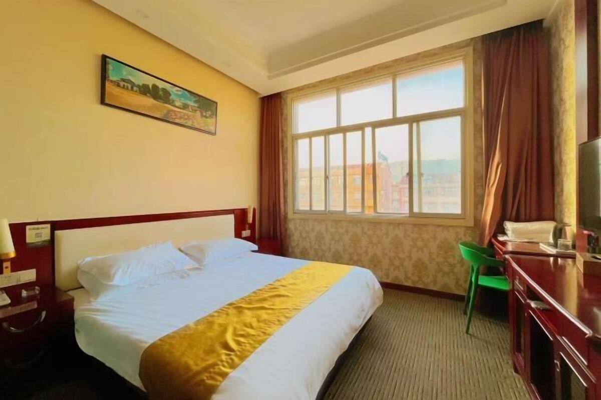 A hotel room with a bed, a desk with a chair, a window with curtains, and a painting on the wall.