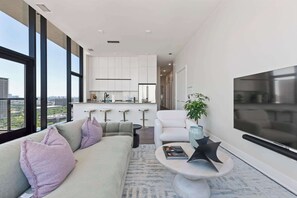 Una sala de estar moderna con un sofá, una mesa de centro y un televisor. Hay una zona de cocina con armarios blancos y un ventanal grande.