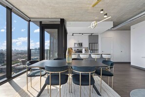 Un comedor moderno con una mesa grande y sillas, una cocina al fondo y vistas a la ciudad a través de amplios ventanales.