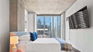 Une chambre d’hôtel moderne dotée d’un grand lit, d’une table de chevet avec une lampe, d’une vue sur la ville grâce à de grandes fenêtres du sol au plafond et d’une œuvre d’art accrochée au mur.