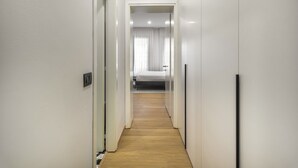 Een smalle gang met een houten vloer die leidt naar een slaapkamer met een bed en een raam met gordijnen.