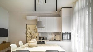 Een moderne keuken met witte kasten, een marmeren werkblad en een ingebouwde oven. Er staan twee witte stoelen bij de eettafel, en een vaas met takken op het aanrecht.