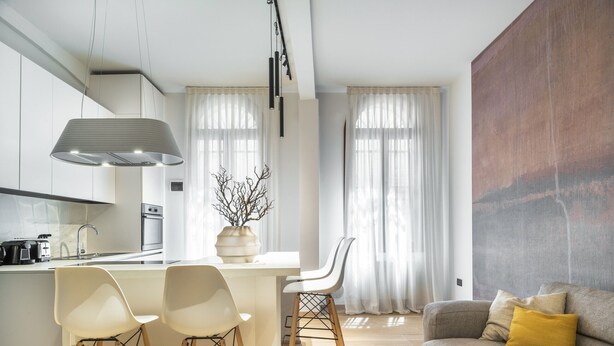 Een moderne keuken met een centraal eiland, twee witte stoelen, een houten eettafel en een grijze bank met een geel kussen.