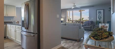 Una cocina moderna con un refrigerador de acero inoxidable, un comedor con mesa de vidrio y una sala de estar con sofá y ventanas amplias.