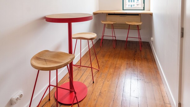 A room with a wooden floor, a red table with stools, a window with blinds, and a white ceiling. - SATURdAYS (Lisbon)