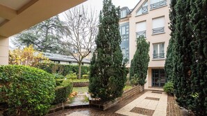 A courtyard with a tiled floor, surrounded by neatly trimmed hedges and tall evergreen trees. - Geranimus Versailles Charost (Versailles)