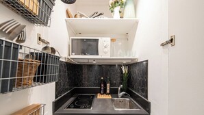 A compact kitchen with a microwave, sink, and stove. There are shelves above the microwave and baskets on the left. - Geranimus Versailles Charost (Versailles)