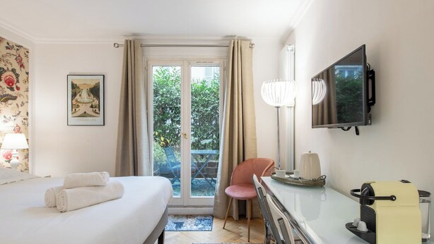 A hotel room with a bed, a desk, a television, and a view of greenery outside. - Geranimus Versailles Charost (Versailles)