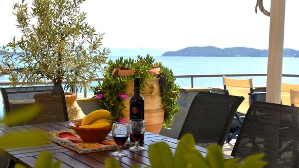 Ein Tisch mit einer Weinflasche, Gläsern, einer Obstschale und einem Blumentopf auf einem Balkon mit Blick aufs Meer.