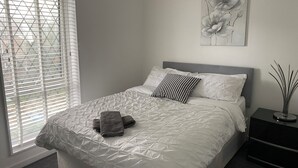 Una cama bien tendida con una cubierta blanca acolchada, un cabecero gris y una almohada de estampado blanco y negro. Una toalla gris está doblada sobre la cama. Un cuadro de flores cuelga de la pared encima de la cama.