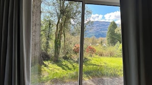 Una habitación con un amplio ventanal corredera que da a una vista panorámica al aire libre: un espacio cubierto de césped, árboles y montañas a lo lejos.