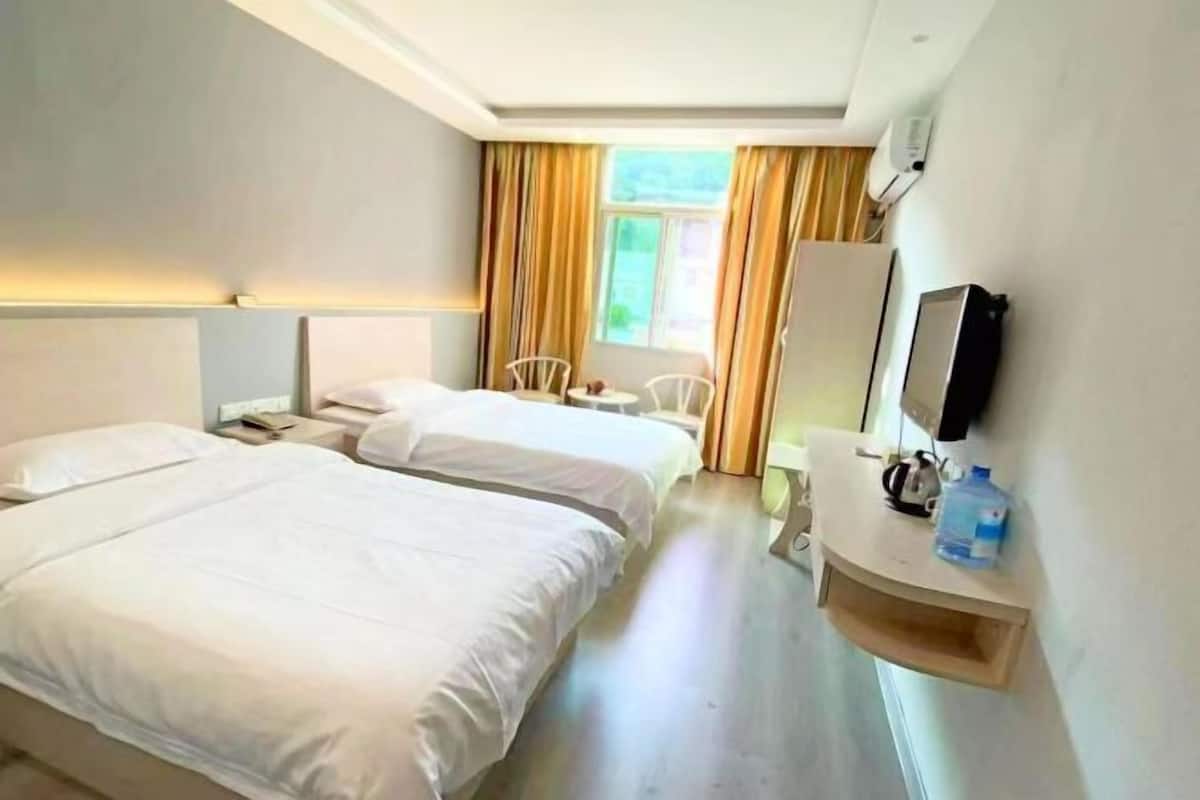 A hotel room with two beds, a small table, a chair, a TV, and a window with curtains.