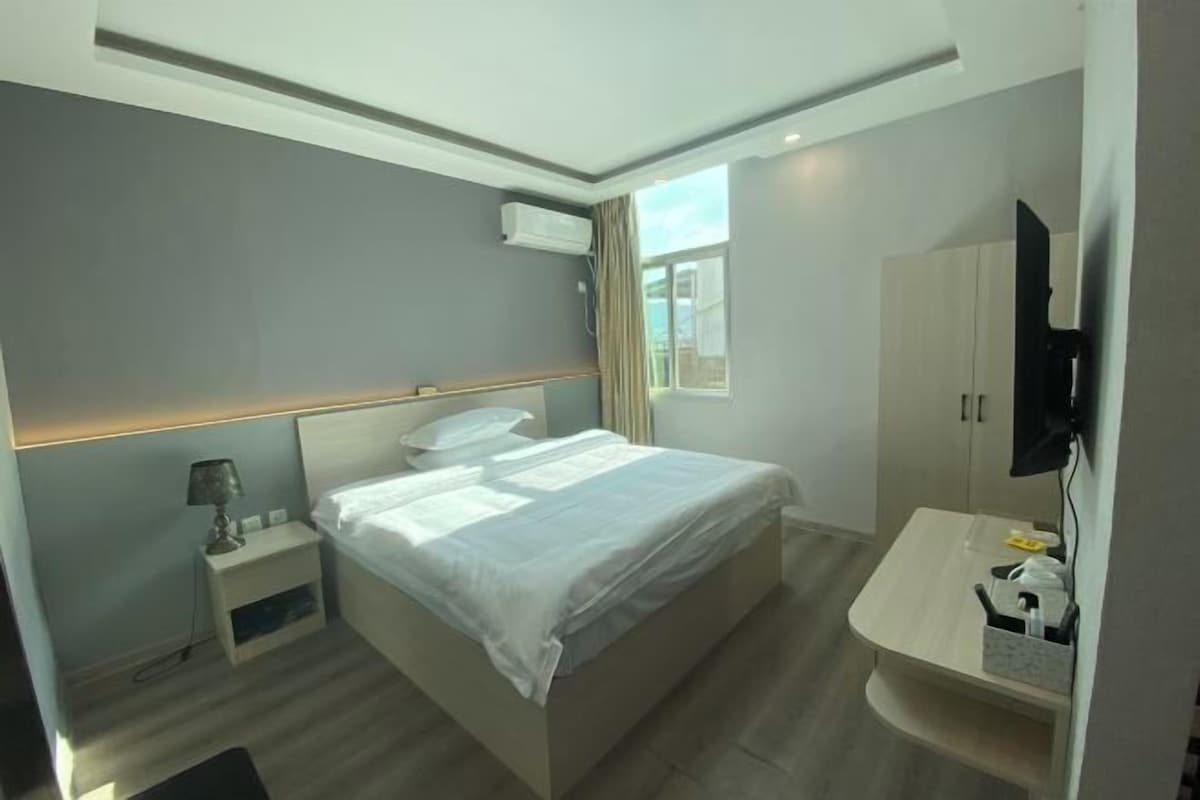 A modern hotel room with a bed, bedside table, lamp, TV, and a small desk with a chair.