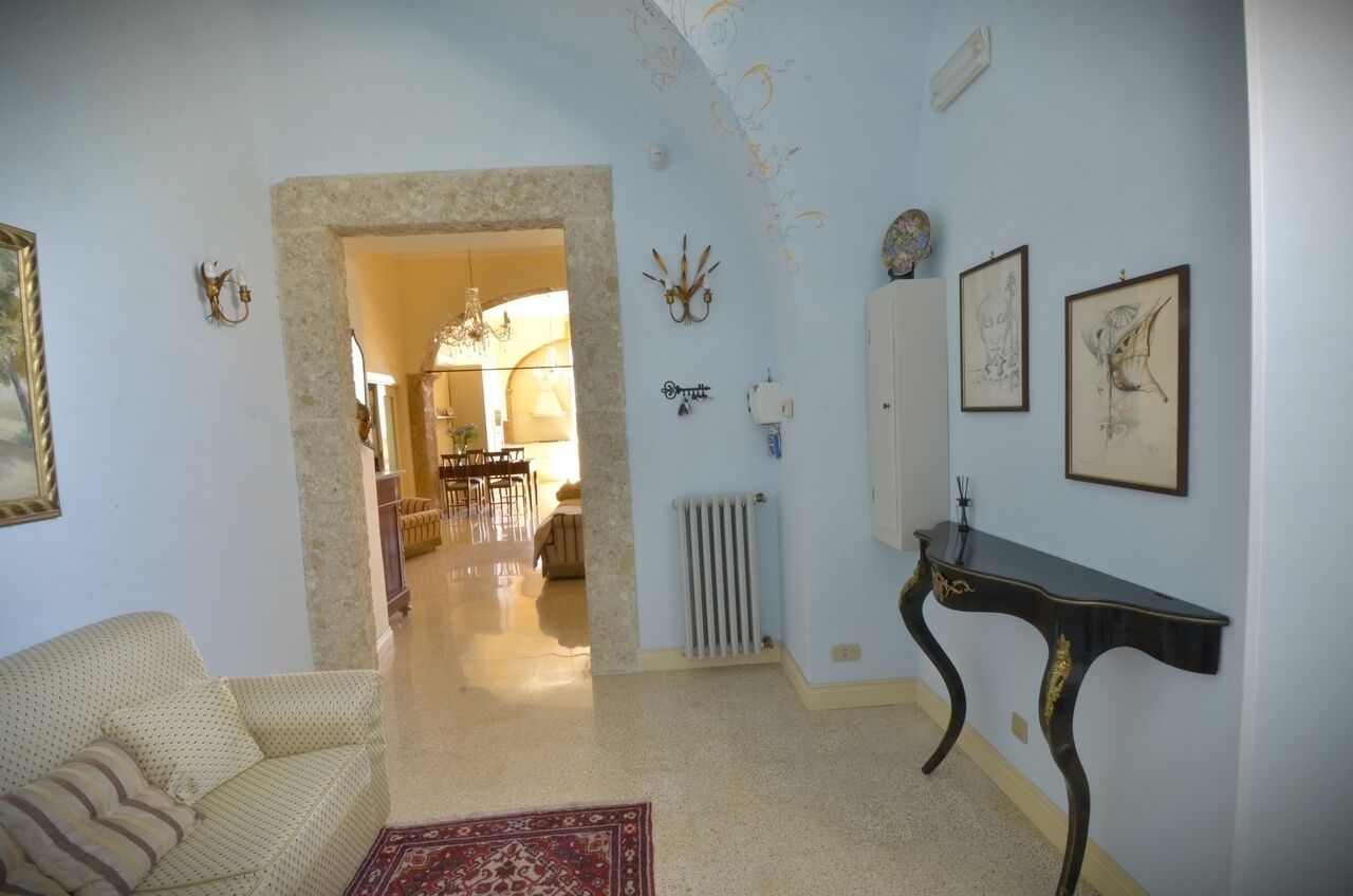 Sala de estar com sofá, mesa e radiador. Um corredor leva a outro cômodo com um lustre.