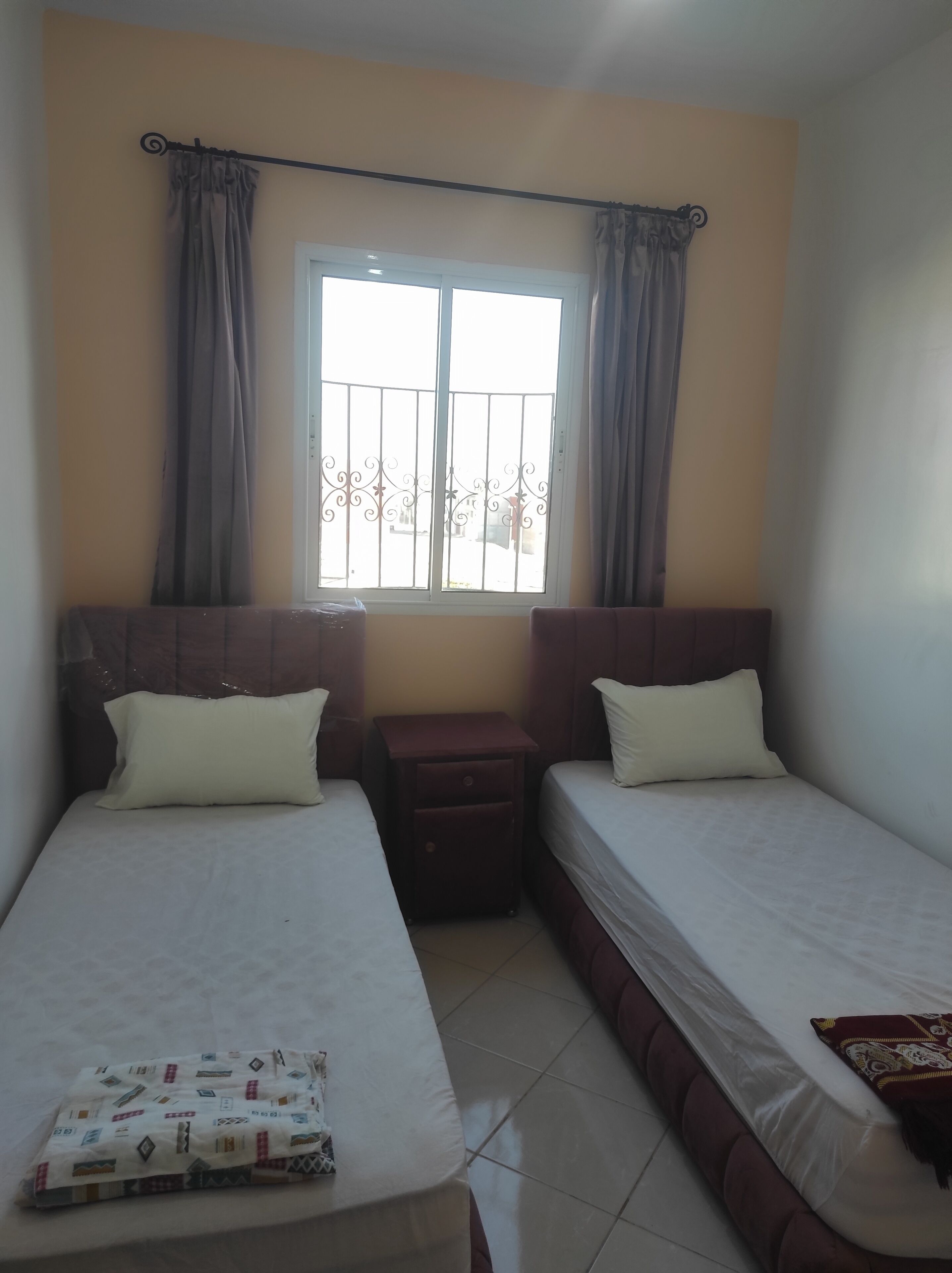 A room with two single beds, a small wooden nightstand, a window with curtains, and a patterned blanket on the floor.