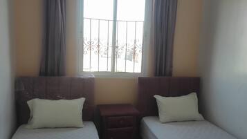 A room with two single beds, a small wooden nightstand, a window with curtains, and a patterned blanket on the floor.