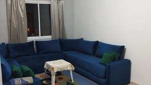 A living room with a blue sectional sofa, a small white table with a floral tablecloth, and a colorful rug. - Apartment 5 minutes from Anza beach (Agadir)