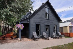 A two-story house with a dark exterior, a front porch with Adirondack chairs, and a hanging flower basket. - Authentic CB, Historic Remodeled Elk Ave Gem! (Crested Butte)