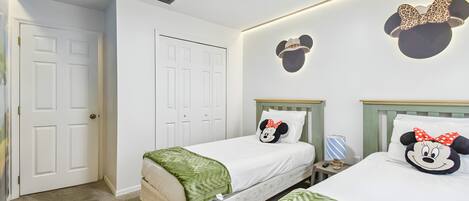 Ett hotellrum med två sängar, var och en med en kudde med motiv av Musse Pigg, en grön pläd och ett nattduksbord med en lampa.