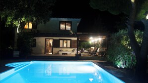 A nighttime view of a house with a swimming pool, a patio with outdoor furniture, and a well-lit entrance. - Villa with pool and basketball court, pet friendly  (Thimari)