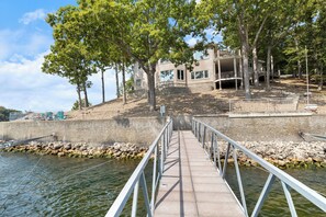 A concrete pier leading to a building with multiple windows and balconies, surrounded by trees and a rocky shoreline. - Pet-Friendly Home In The Main Channel w/ Breathtaking Sunsets & Lake Views (Lake Ozark)