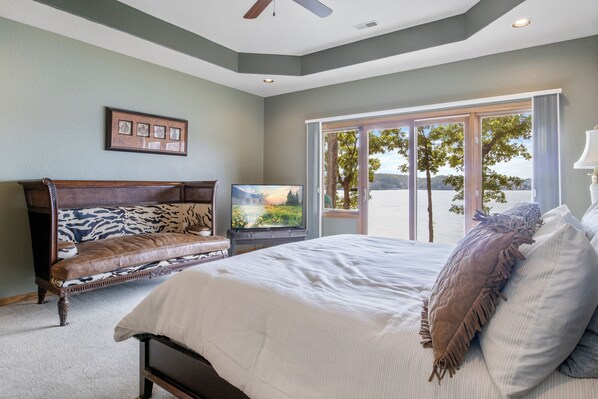 A bedroom with a large bed, a TV, a bench, and a view of the outdoors. - Pet-Friendly Home In The Main Channel w/ Breathtaking Sunsets & Lake Views (Lake Ozark)