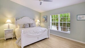 Un dormitorio con cama blanca, dos mesitas de noche con lámparas, ventilador de techo, ventana con persianas y obras de arte en las paredes.