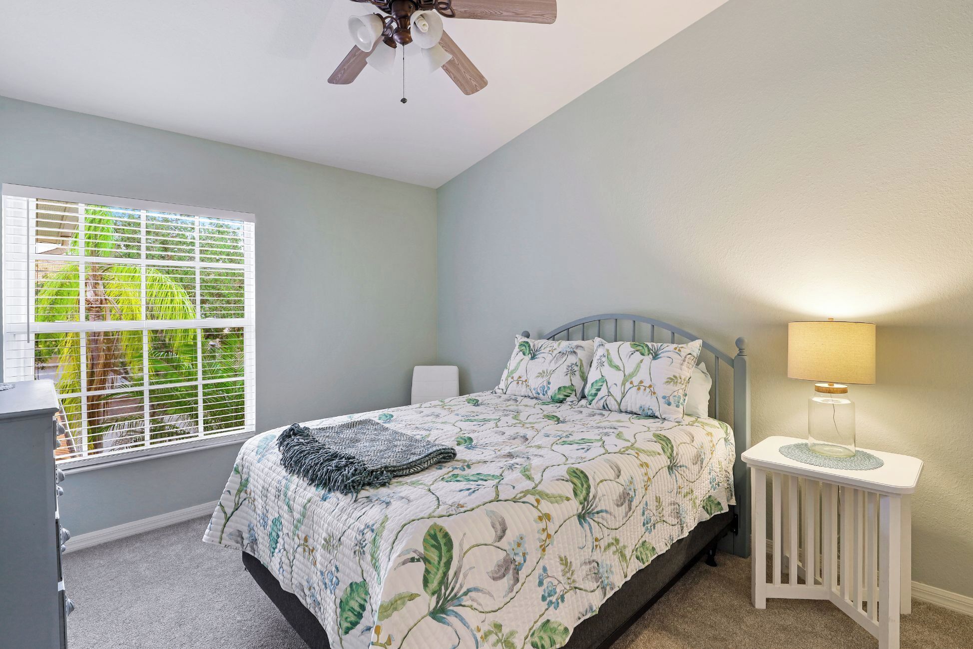 A bedroom with a bed, a nightstand, a lamp, a ceiling fan, and a window with blinds.