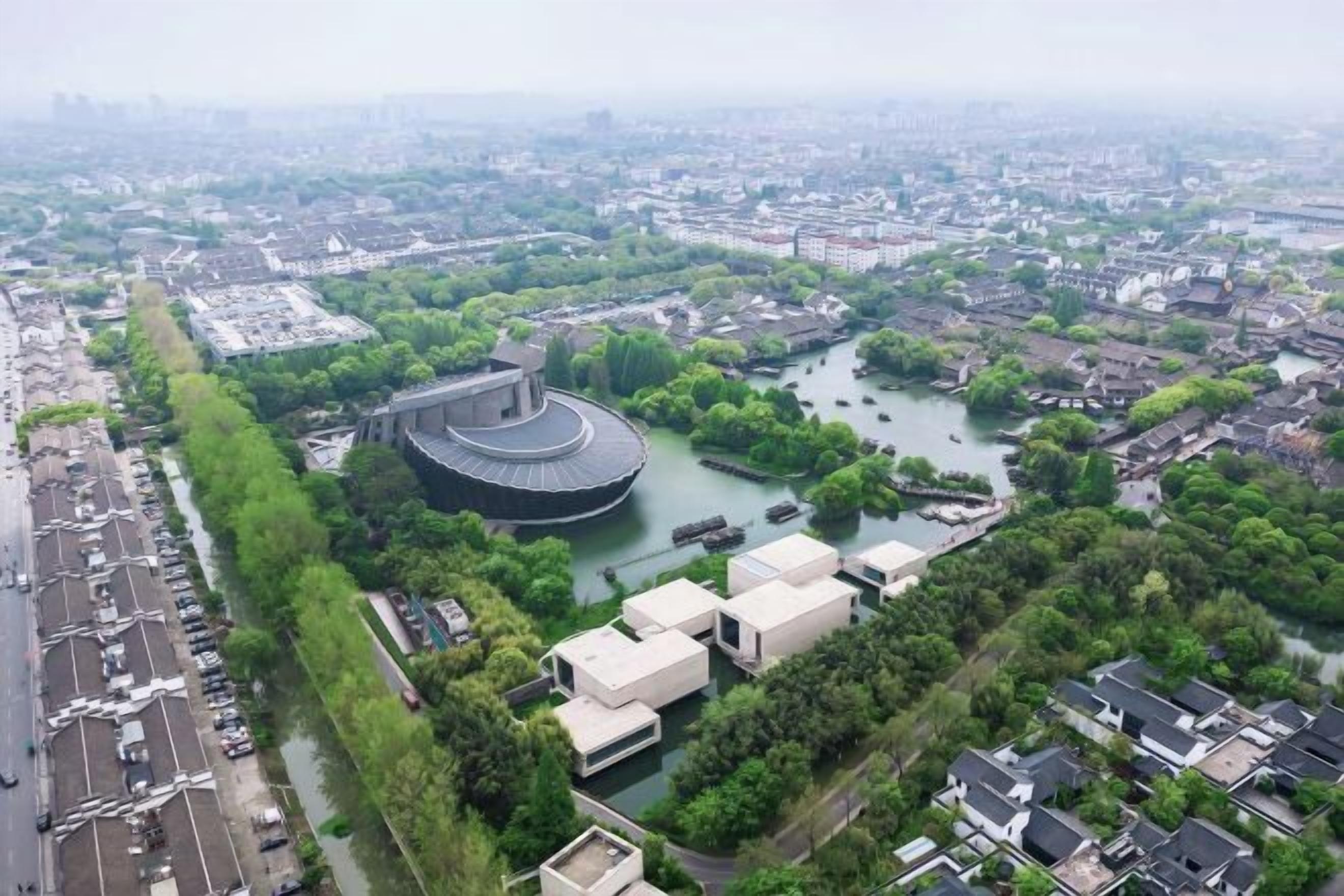 Vista aérea de una ciudad con un gran edificio circular rodeado de agua, vegetación y zonas residenciales.
