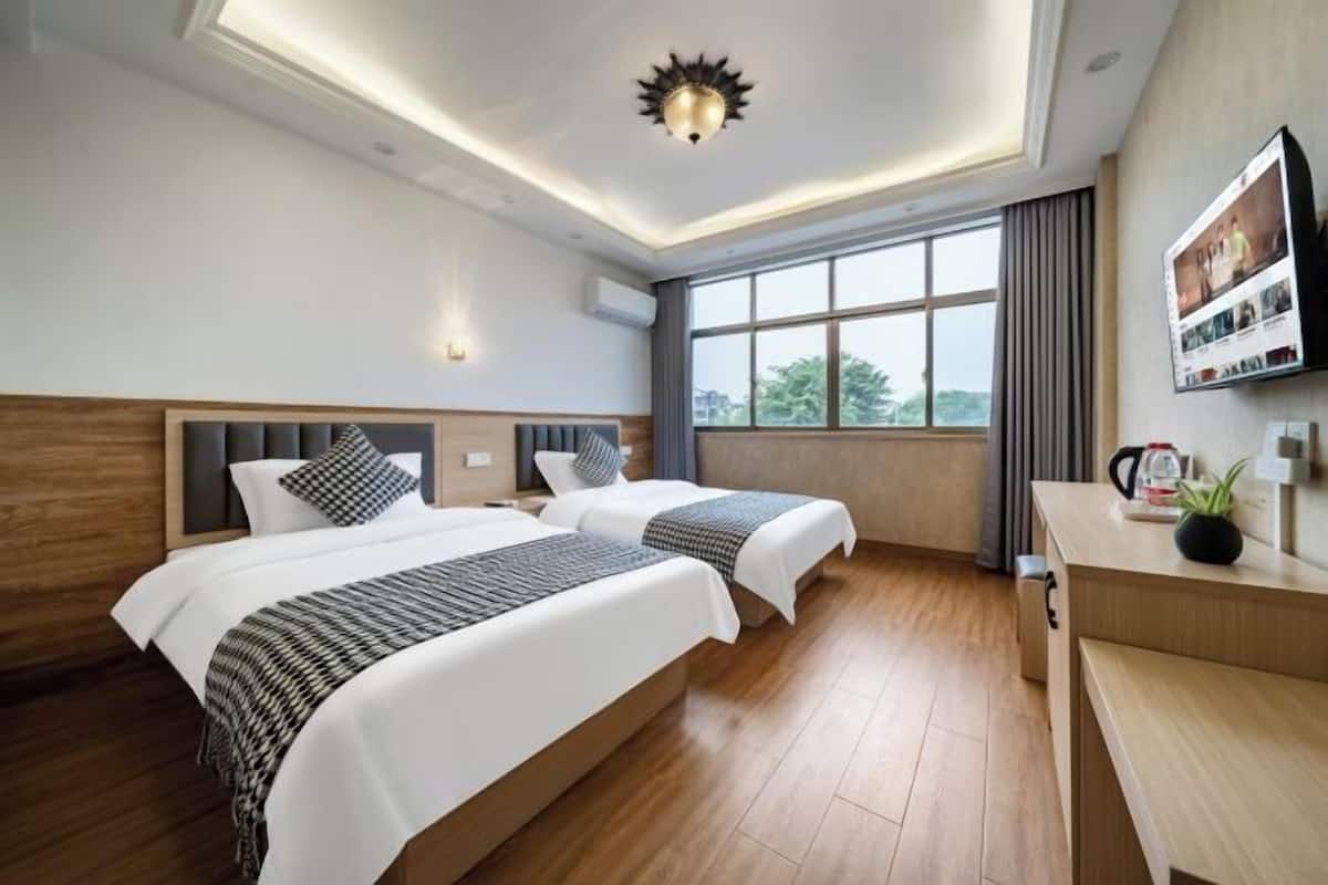 Ein Hotelzimmer mit zwei Betten, Holzboden, großem Fenster mit Vorhängen, Schreibtisch mit Pflanze und Wand-Fernseher.