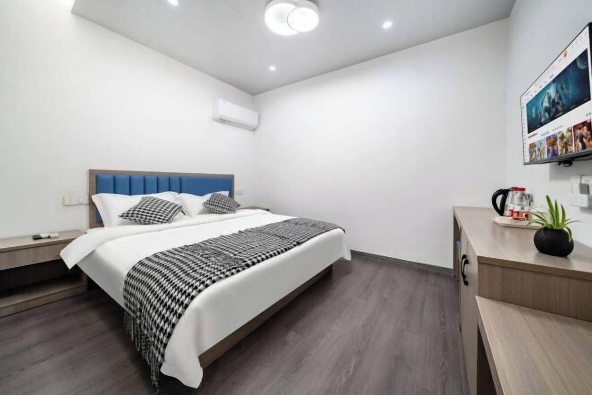Ein modernes Hotelzimmer mit Bett, Nachttischen, Schreibtisch, Stuhl, Zimmerpflanze und einem wandmontierten Fernseher.