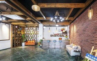 Amplia habitación con techo alto, vigas de madera a la vista, pared de ladrillo y una zona de estar con sofá y taburetes de bar.