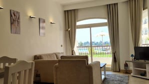 Ein Wohnzimmer mit Sofa, Sessel und TV-Schrank. Ein großes Fenster mit Vorhängen, und draußen ist ein Balkon mit Geländer zu sehen.