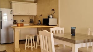 Eine kompakte Küche mit weißen Schränken, einem Kühlschrank, einer Mikrowelle und einer Kaffeemaschine. Auf dem Holztisch stehen zwei weiße Stühle und eine blaue Vase.