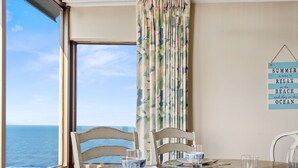 Un comedor con vista al océano, una mesa dispuesta con platos, tazones y vasos, y un letrero en la pared que dice "SUMMER a time to RELAX at the BEACH and play in the OCEAN".