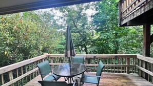 Een houten terras met een glazen tafel en stoelen, omgeven door weelderige begroeiing.