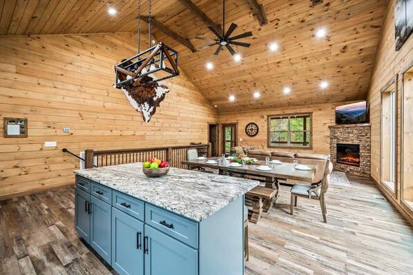 A spacious cabin interior with a wooden ceiling, a stone fireplace, a dining area, and a kitchen with blue cabinets and a marble countertop. - Luxe 4BR w/Firepit*Hot Tub*Game Room*Comm Pool (Sevierville)