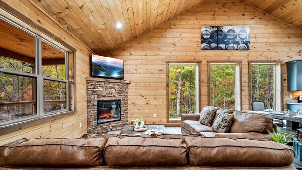 A cozy cabin interior with a stone fireplace, leather sofa, and large windows offering views of the forest.