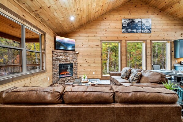 A cozy cabin interior with a stone fireplace, leather sofa, and large windows offering views of the forest.